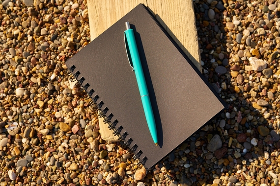 A black spiral notebook and blue pen sit on a blank of wood resting on some gravel