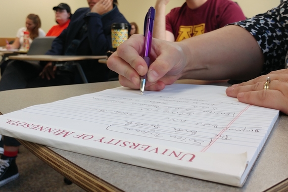 closeup of hand writing on a University of Minnesota notepad in the foreground of a classroom where other students are seated in a candid style