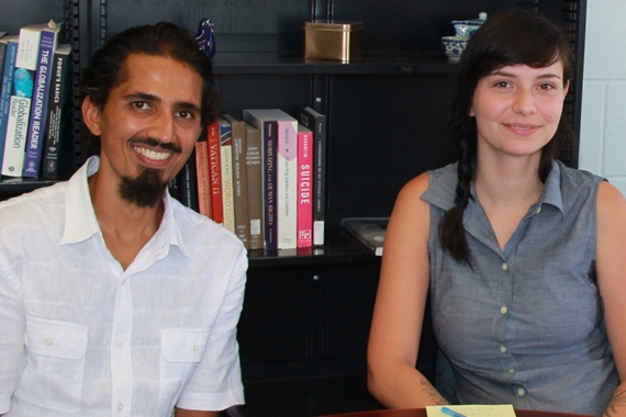 Photo of 2 graduate students, male and female, sitting in front of a bookshelf. They are smiling at the camera.