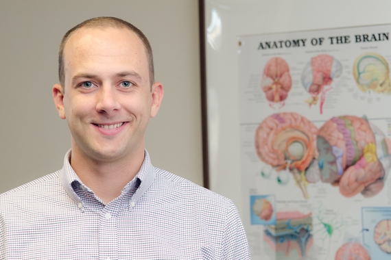 Male graduate student looking at the camera and smiling. A poster with a diagram of a brain is on the wall behind him.