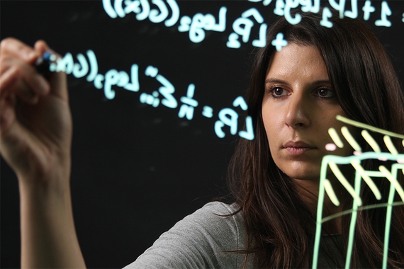 Sara Algeri draws a statistical formula on a large piece of glass with colorful markers. She is behind the glass.