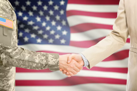 two arms shaking hands in front of an American flag, one arm dressed in military attire, one dressed in a tan suit