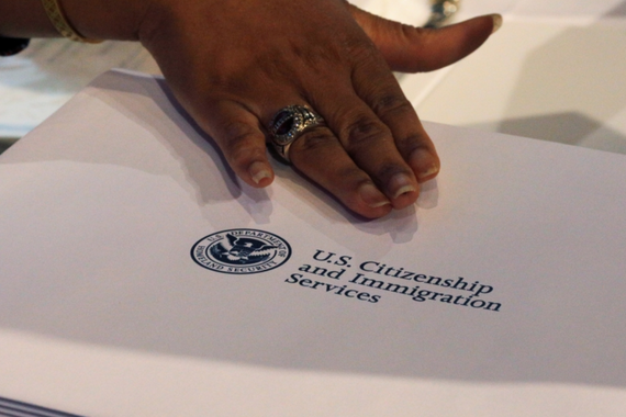 A hand pushing a stack of papers that say "U.S. Citizenship and Immigration Services" with the US Department of Homeland Security symbol