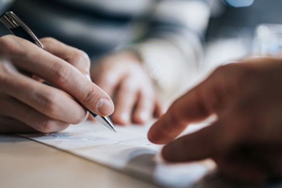 a person's hand holding a pen close to a paper, writing, while another person's hand points at the paper