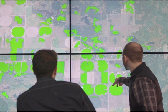 Eric Shook and Jesse Bakker look at a large computer screen that is showing cropland highlighted in bright green.