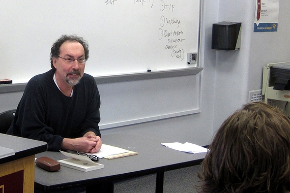 Professor Emeritus Geoffrey Sirc at table in classroom smiling at students (visible is one student's head from the back)