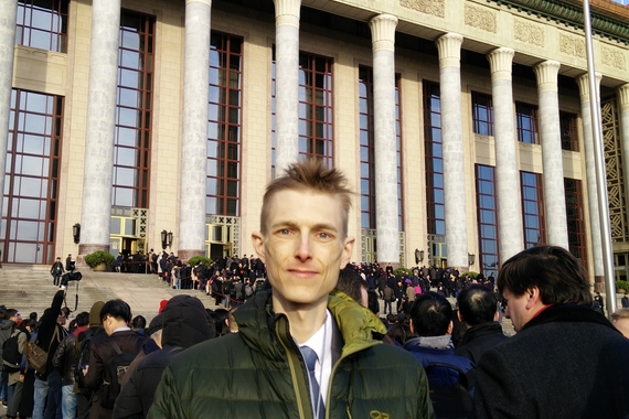 Man standing in front of the Great Hall of the People in Beijing, with crowd gathered on steps.