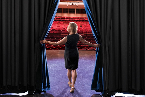 Nancy Kelly parting the curtains on a theatrical stage, facing an audience of empty seats