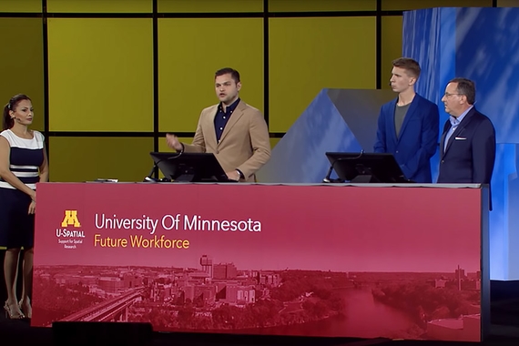 Four people from the Department of Geography, Environment & Society on stage at the Ersi Conference standing behind a U of M banner. One of them is speaking.