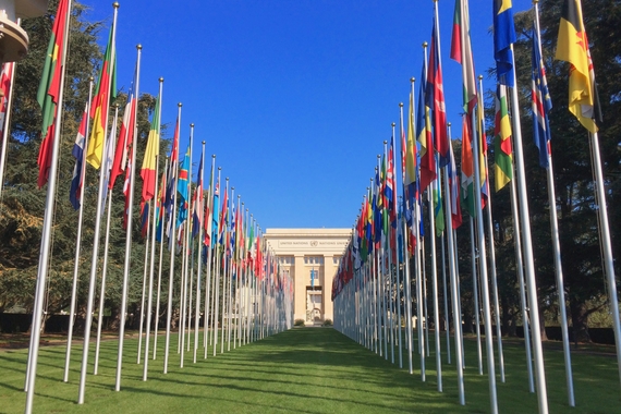 Two straight lines of flag poles displaying United Nations member state flags extending up a grassy alley to the Palais des Nations