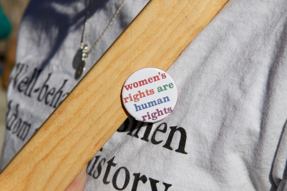 A small, white, circular button that reads "Women's Rights are Human Rights" in multi colored letters sits on top of the chest of someone wearing a grey tee-shirt with a brown bag strap across their body.