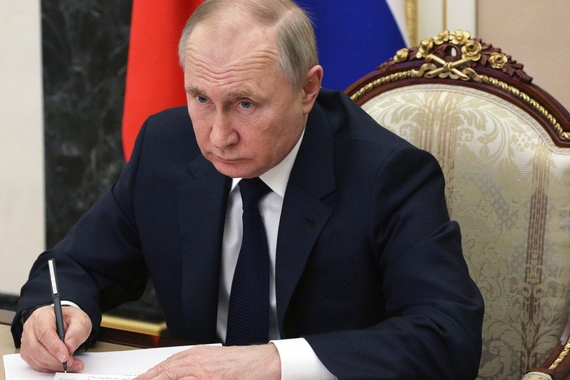 Vladimir Putin sitting in a chair, signing a sheet of paper with a pen
