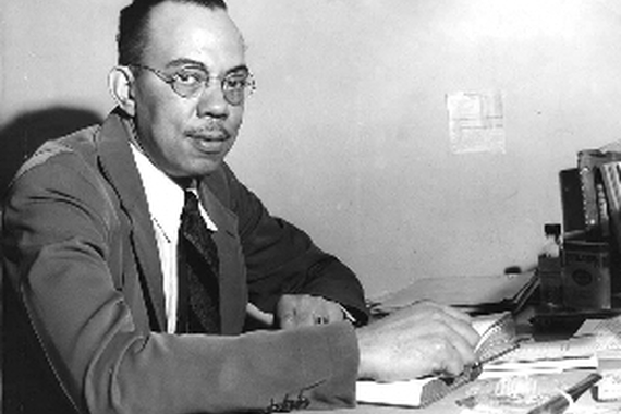 A man wearing a suit and glasses looks up from the book he is reading at a desk