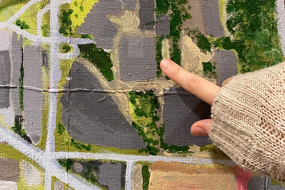 Someone points to a spot on a hand-painted map, a composition of gray buildings, light gray roads, and green trees