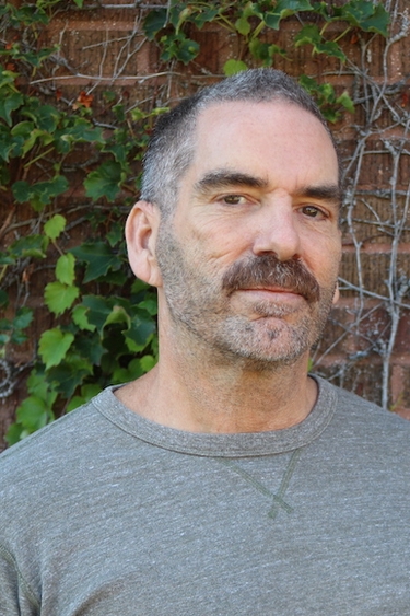 Kevin Murphy looking into the camera with a serious expression in front of a brick building with vines