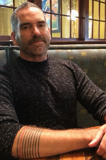Kevin Murphy, a person with short grey hair and a mustache with a tattoo showing on their forearm