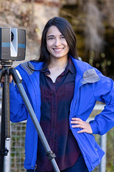 Samantha Thi Porter, a person with light skin and long, black hair wearing a bright blue jacket and dark top. Samatha stands next to a surveying device on a tripod. There is a brick wall and a metal gate in the background.