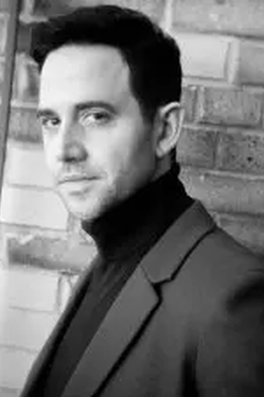 Santino Fontana in turtleneck and blazer stands in front of brick wall.