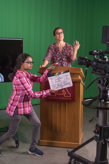 A young girl holds a clapperboard and an adult woman speaks behind a podium, both recorded by a film camera