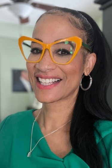 A woman with funky orange glasses and a long black ponytail smiles at the camera
