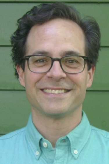 A white man with brown hair wearing glasses and a teal button-up shirt smiles at the camera