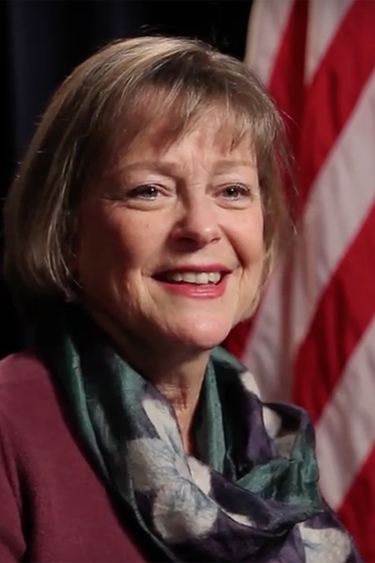 Kathleen Kuehnast, a person with light skin and light brown bobbed hair, wearing a blazer and a scarf. A flag of the United States is in the background.
