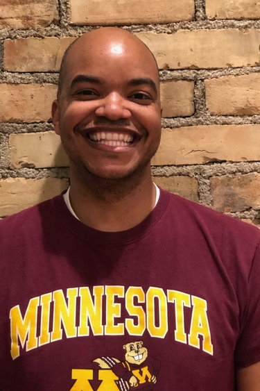Rod Lature, a person with brown skin and no hair, wearing a University of Minnesota shirt and smiling