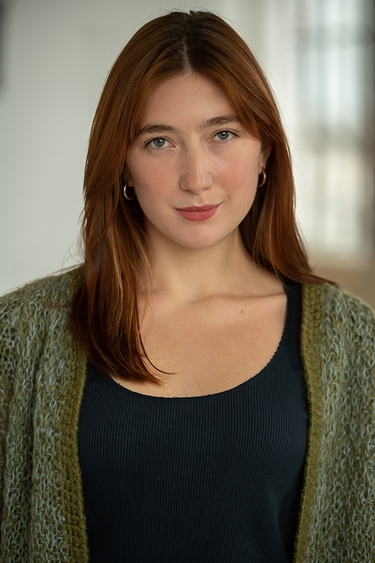 Female Actor wearing a black top with green shall, looking into camera confidently.