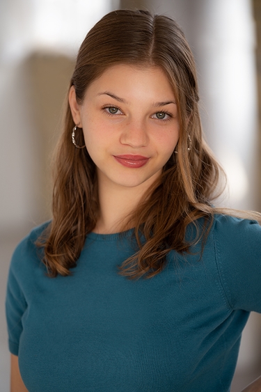 Female Actor wearing a teal shirt looking into camera confidently with a slight smile.