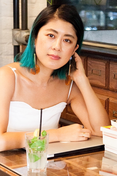 Person with dark brown and turquoise hair to shoulders and light skin seen from waist up, wearing thin strapped white top and sitting at a table on wood bench; drink and stack of books on table