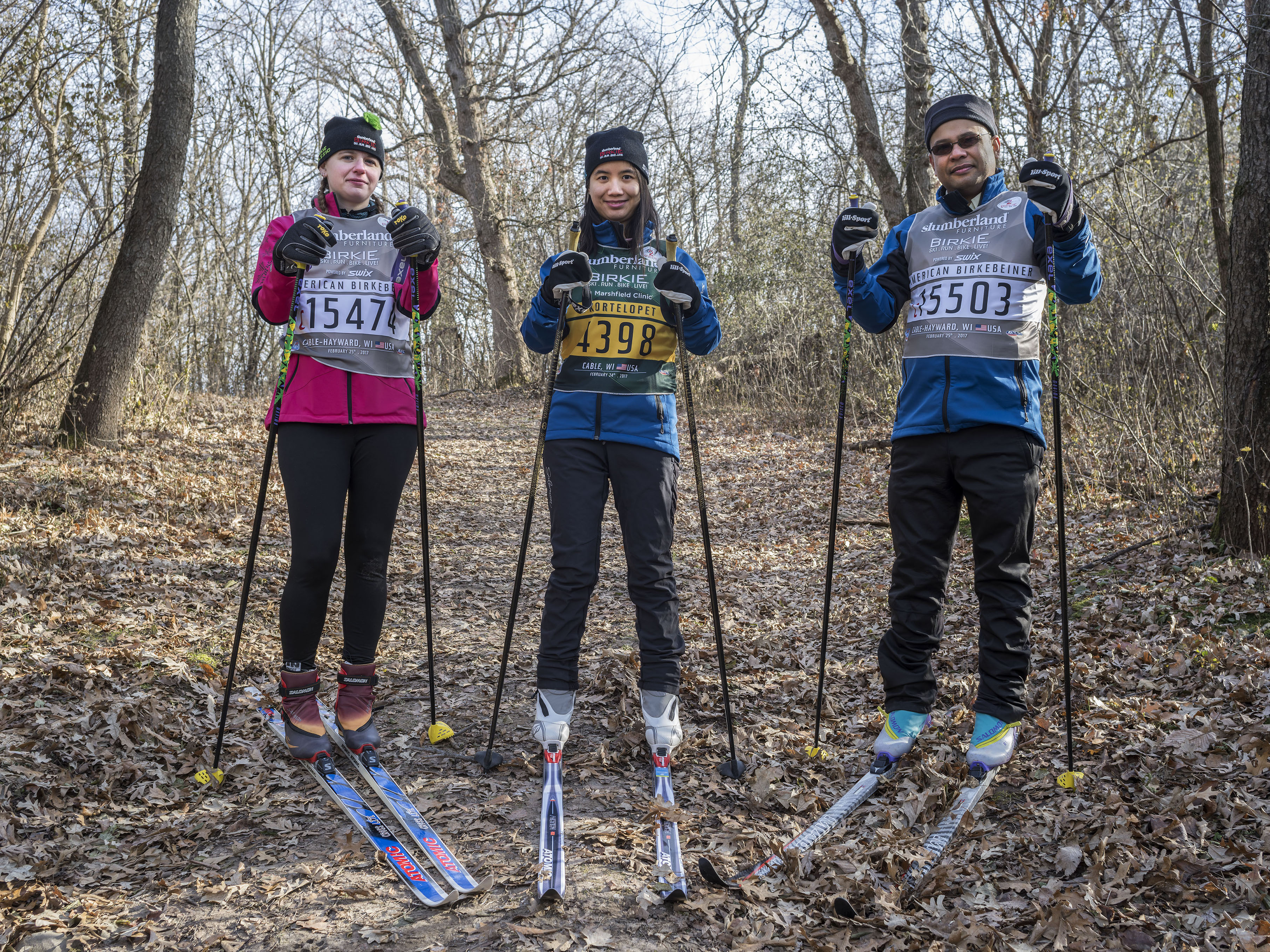 Three cross country skiers stand in a snowless woods dressed in race bibs 