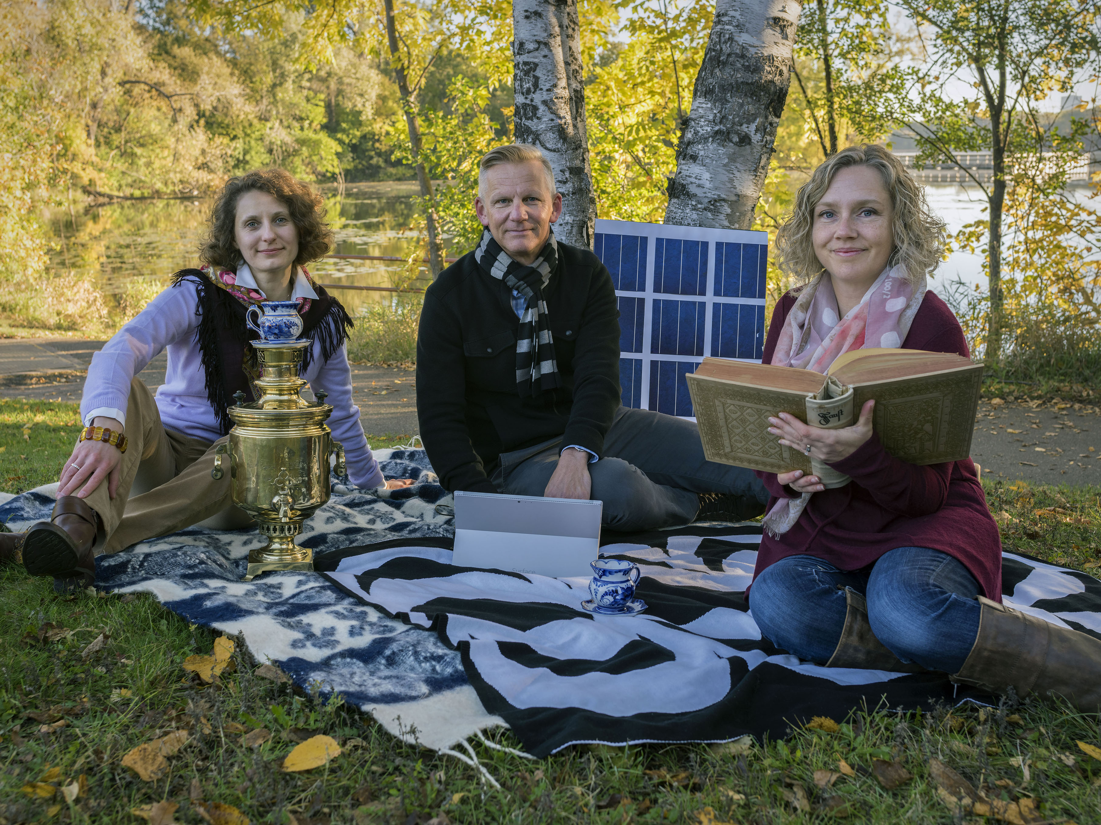 Three instructors sit in the grass at cedar lake with a antique Russian tea set, solar cells, and a antique book.