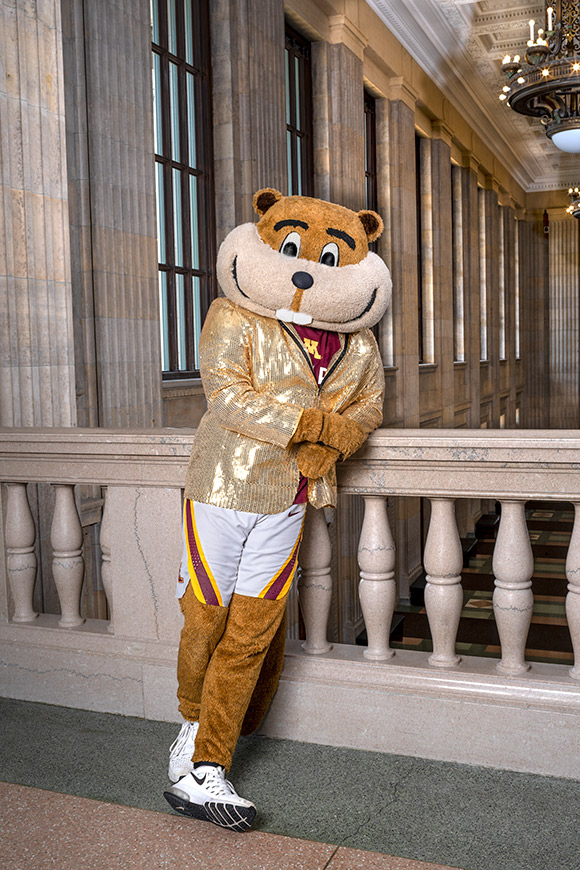 Photo of University of Minnesota mascot Goldy Gopher wearing a sparkly jacket leaning against a bannister in side Northrop auditorium