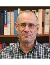 A man with glasses and a plaid shirt is pictured from the shoulders up in front of books
