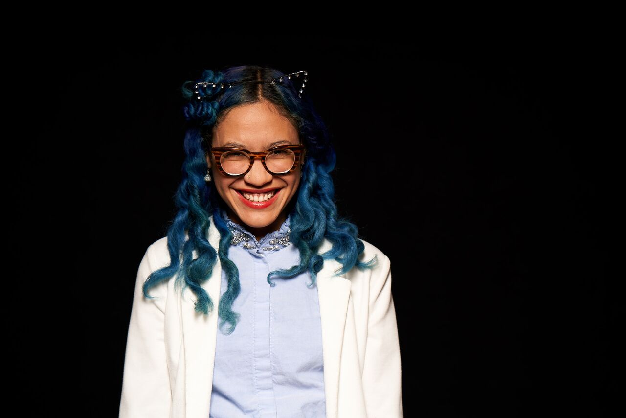 smiling african american woman with blue hair and glasses stands against a black background