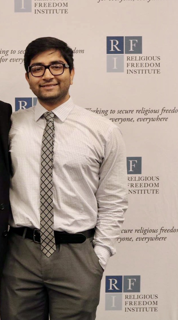 A young man with black glasses wearing a white button up, grey and blue patterned tie, and grey slacks stands in front of a backdrop with a repeating pattern reading "RFI Religious Freedom Institute."