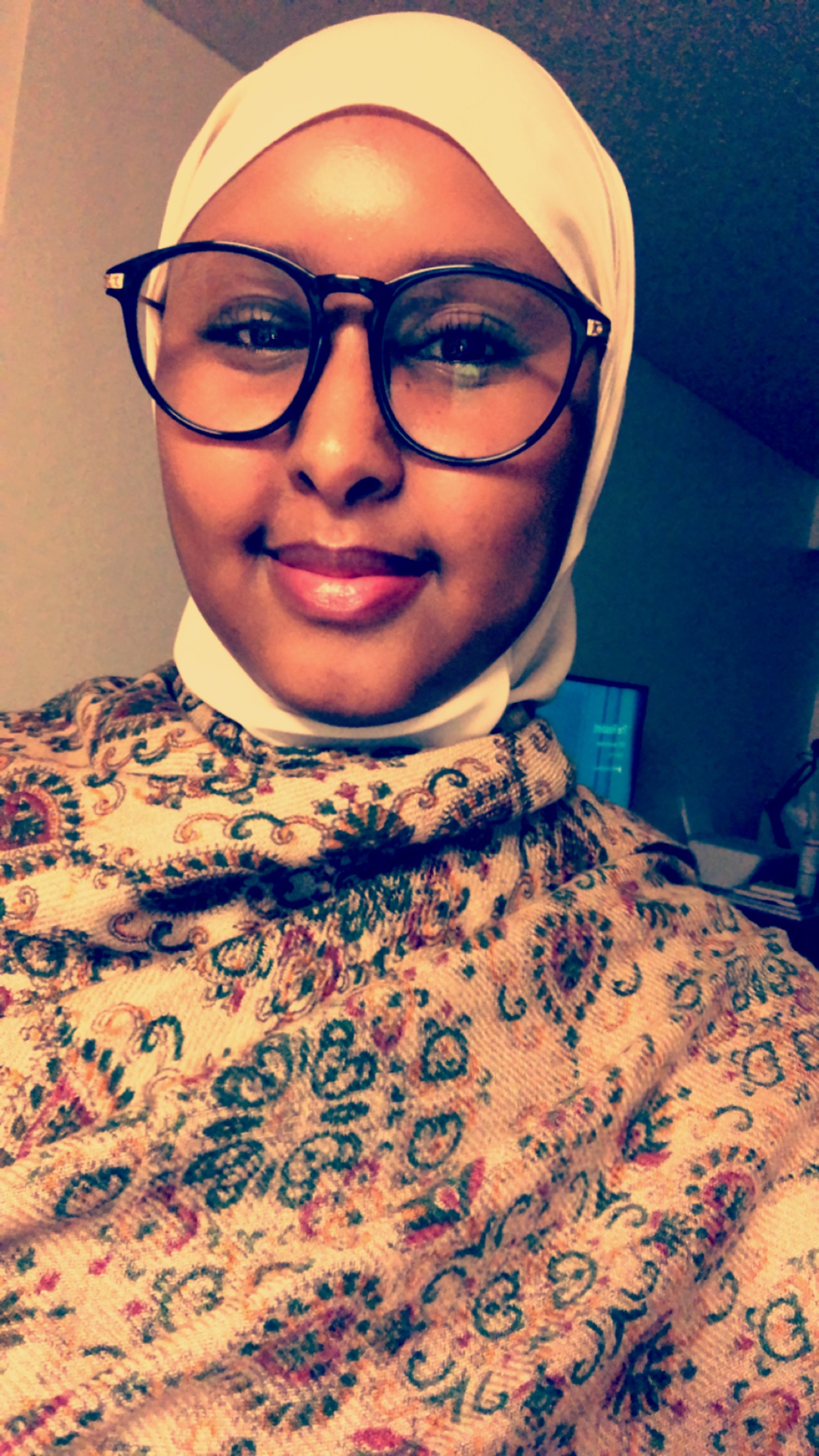 A photo of a young woman wearing a floral print top, black glasses, and a white hijab from the shoulders up 