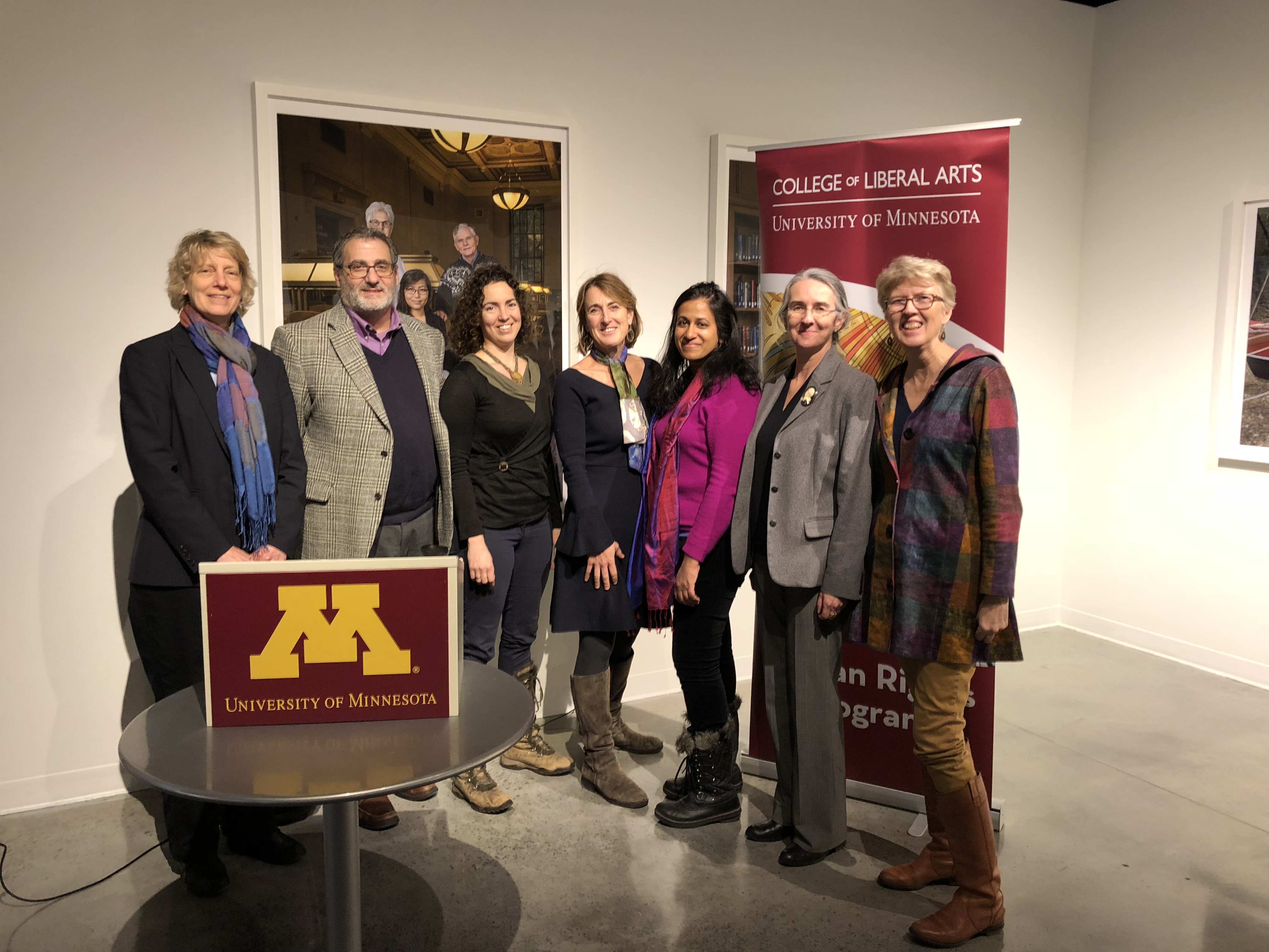 A group of seven adults stand in a line in front of a sign reading "College of Liberal Arts." There are 6 women and one man, and a podium reading "University of Minnesota" is placed on a table in the foreground.