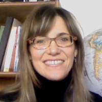 A photo of a middle-age woman's face. She has long light brown hair and side swept bangs and is wearing brown glasses and a black turtleneck top. There is a book shelf with books and a globe behind her.