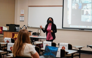 Fahima Aziz wearing a mask talking in a classroom. One person sits in the classroom listening. Projected on a screen behind Aziz is a group of who students tuning into her class through Zoom. 