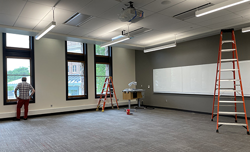 Photo of classroom with light grey carpet, dark grey wall with white board, white ceiling, and three windows with man staring outside one; red step ladder to right side