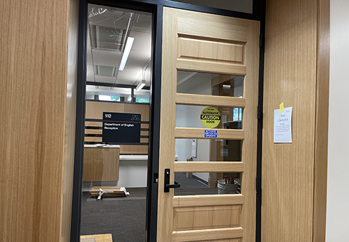 Photo of doorway with wood frame and wood door and dark sign on glass with white lettering 112 Department of English Reception