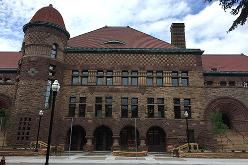 Photo of outside of Pillsbury Hall, yellow sandstone building with red roof and turret, and stone plaza entry