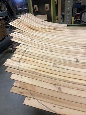 Photo of a sculpture in progress composed of wood strips curving upward on the right side with a curved line drawn across the strips