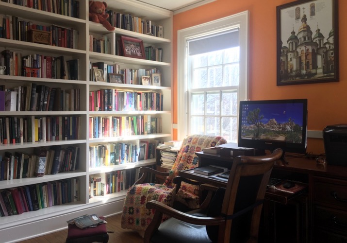 Photo of Prof John Watkins' office, with wall of bookshelves, crochet blanket over rocker with stool, monitor on wood desk