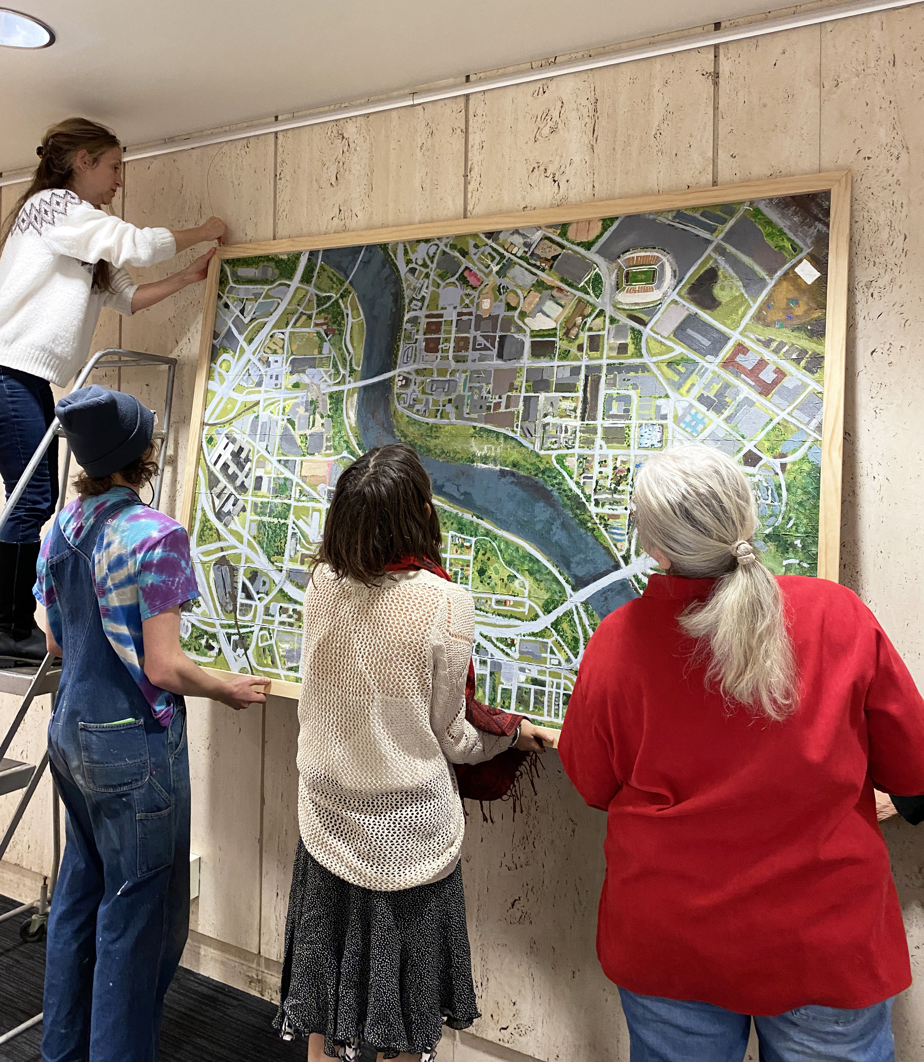 Three people hold a large framed map while another on a step-ladder attaches it to hangers